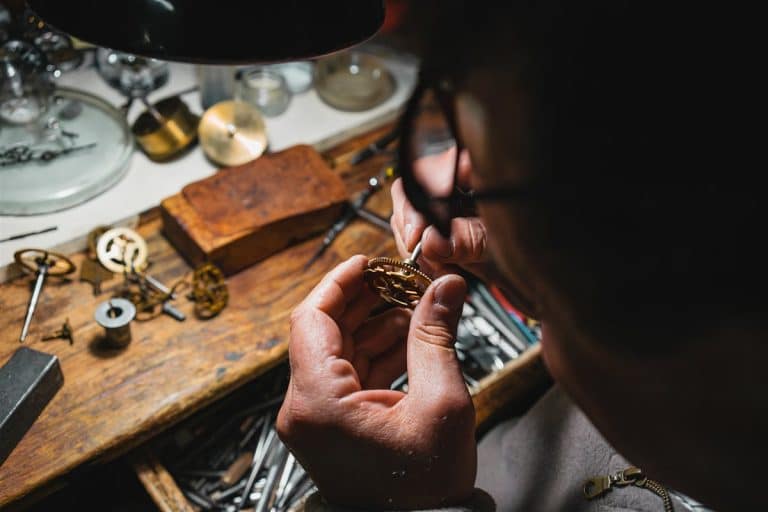 A horologist examining a small gold watch gear at a cluttered workbench