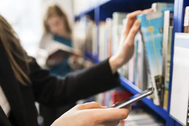 A hand pulling a book from a library shelf, a phone in the other hand