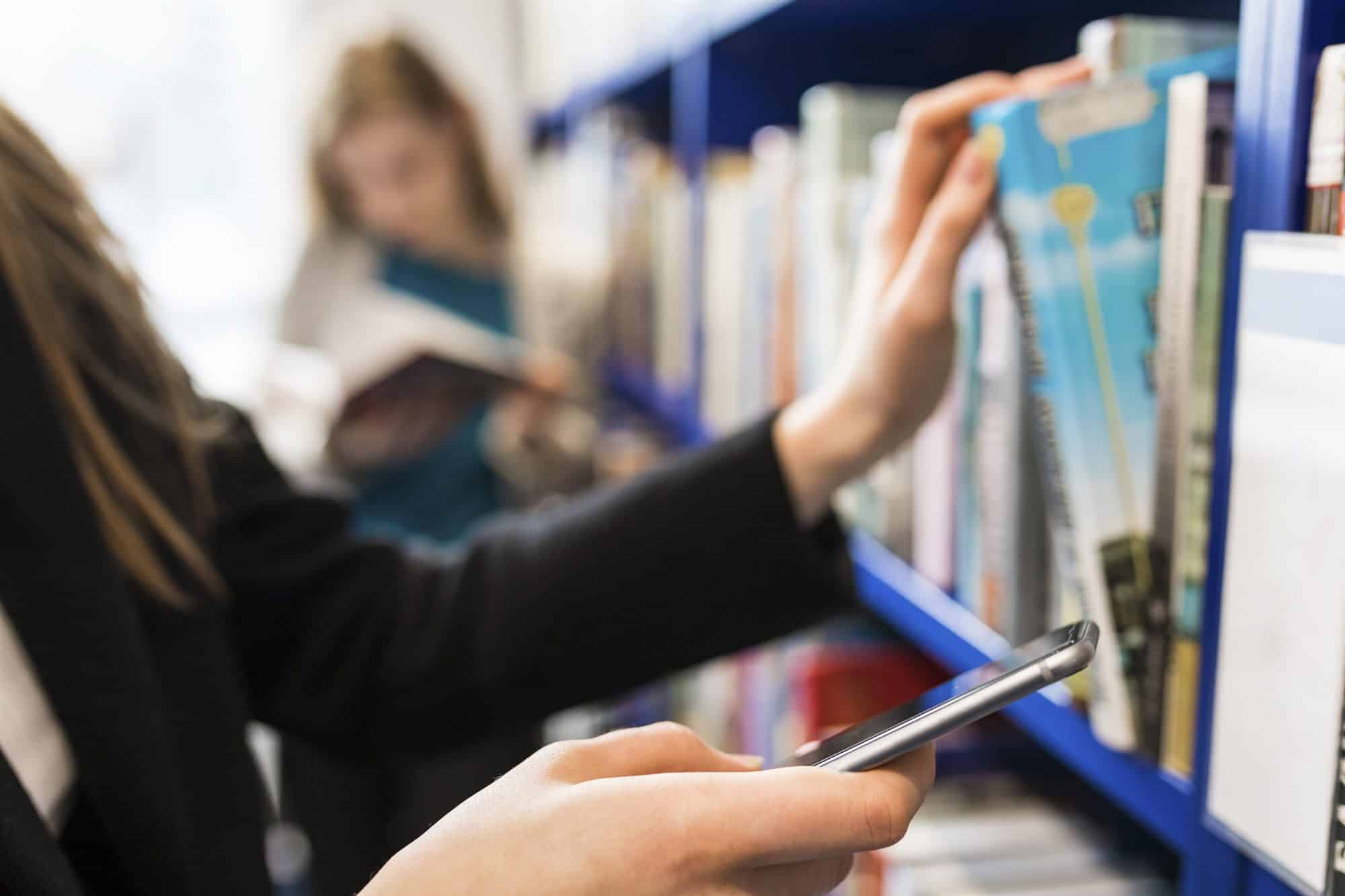 A hand pulling a book from a library shelf, a phone in the other hand