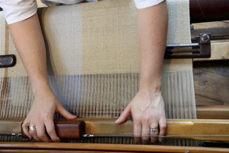 A weaver's hands guiding cream fabric through a traditional wooden loom