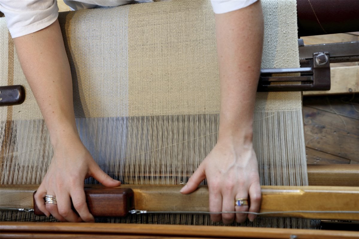 A weaver's hands guiding cream fabric through a traditional wooden loom