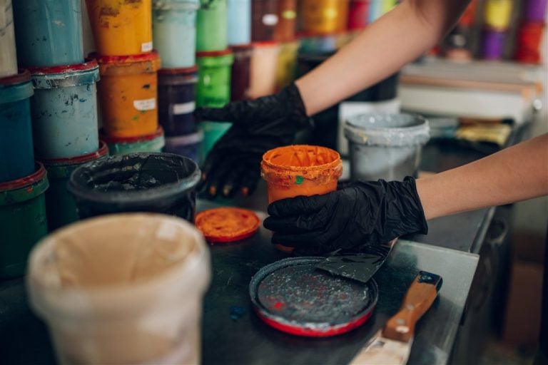 Gloved hands opening a pot of orange paint among stacks of colourful tins in a print workshop