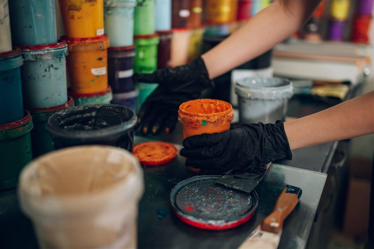 Gloved hands opening a pot of orange paint among stacks of colourful tins in a print workshop