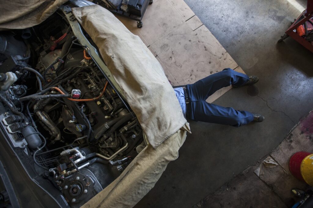 'A mechanic lying under the front of a car in a workshop, with the engine bay stripped open and covered by a dust sheet.'