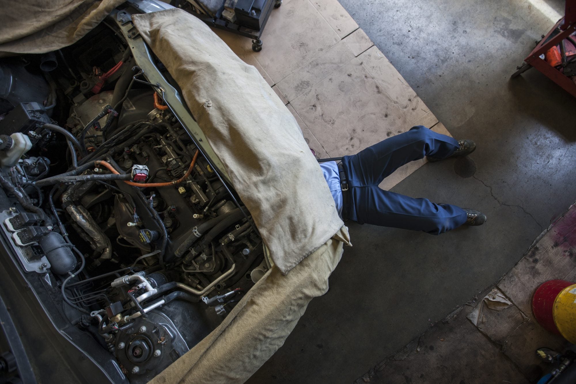 'A mechanic lying under the front of a car in a workshop, with the engine bay stripped open and covered by a dust sheet.'