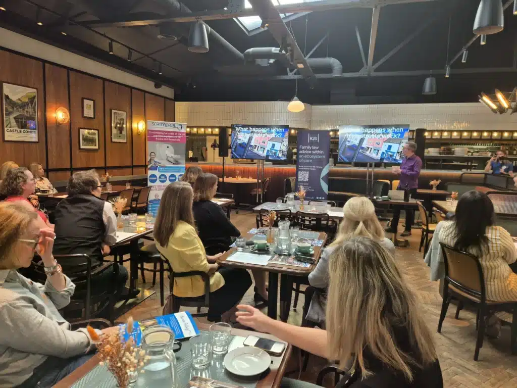A speaker addressing seated guests at a business breakfast, with two screens behind reading What happens when high streets work