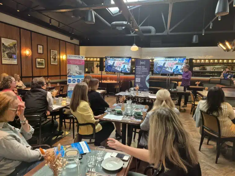 A speaker addressing seated guests at a business breakfast, with two screens behind reading What happens when high streets work