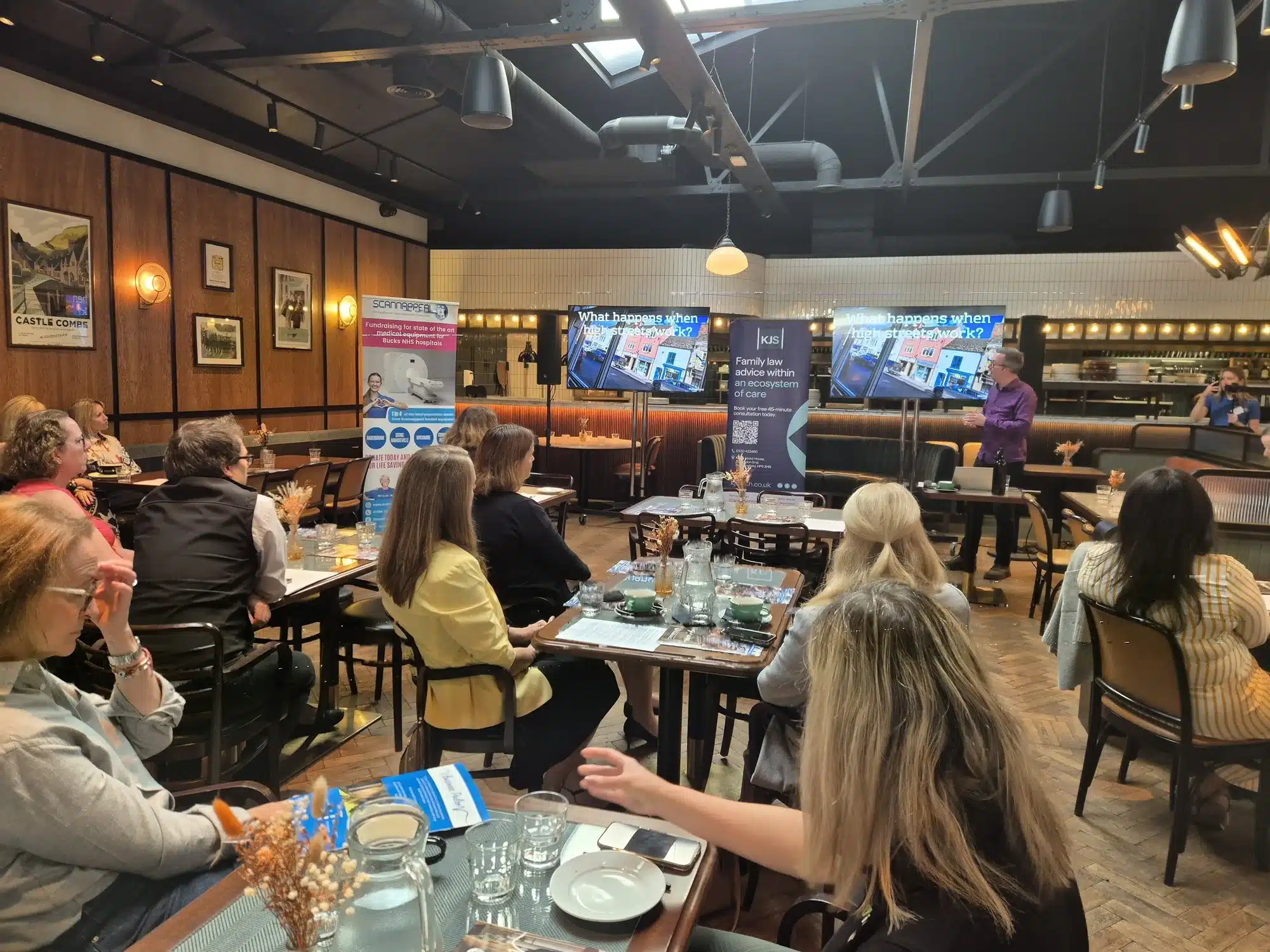 A speaker addressing seated guests at a business breakfast, with two screens behind reading What happens when high streets work